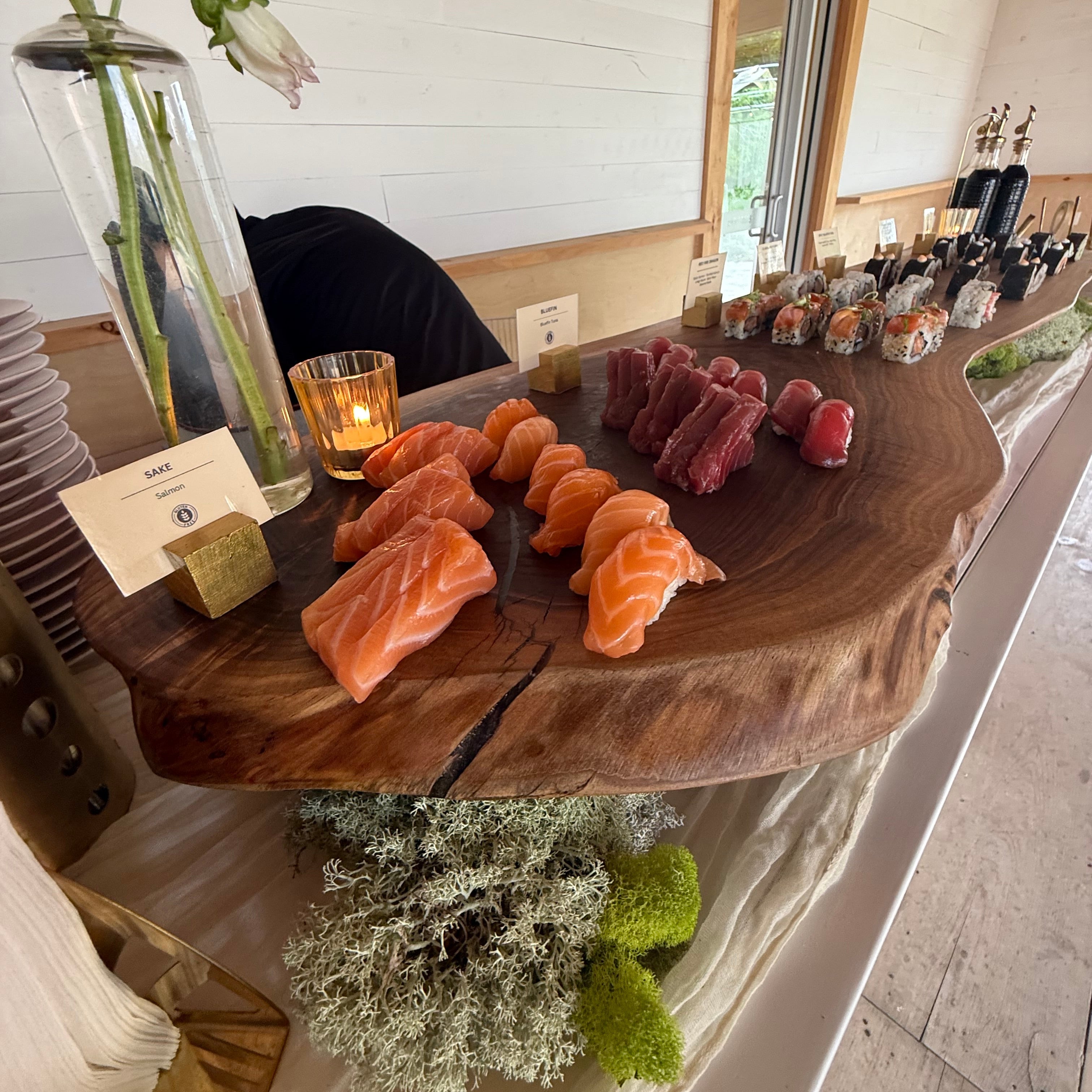Wooden platter with various types of sushi on a white surface with a mirror reflection.